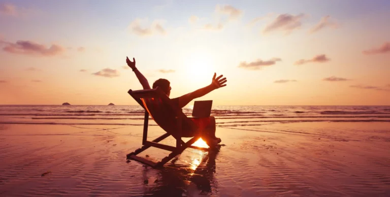 A person sits on a beach chair with a laptop, arms outstretched in joy, facing the sunset over the ocean—perfect inspiration for any Demo Blog. The colorful sky and wet sand reflect the warm light, creating a peaceful scene.