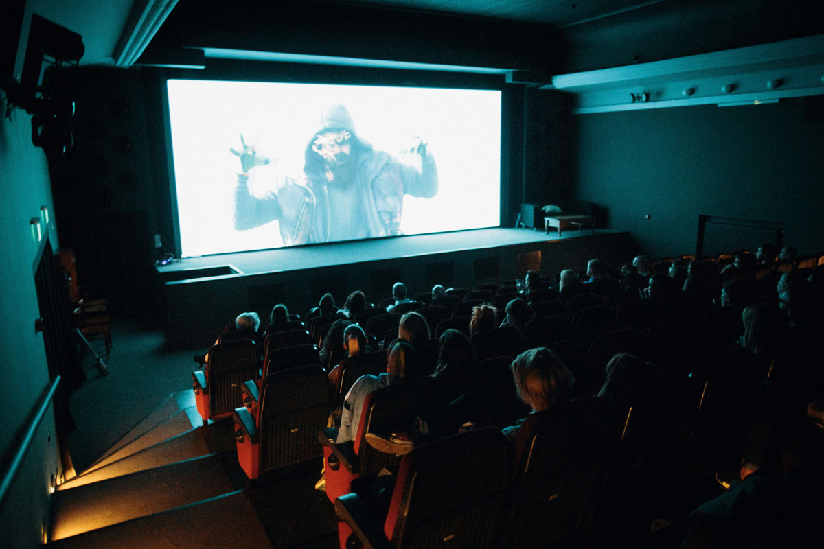 An engaged film audience sits in a dark theater, watching as the screen reveals a person in a hooded jacket with hands raised, illuminated by blue light.