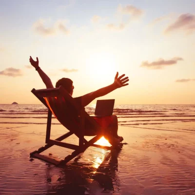 A person sits on a beach chair with a laptop, arms outstretched in joy, facing the sunset over the ocean—perfect inspiration for any Demo Blog. The colorful sky and wet sand reflect the warm light, creating a peaceful scene.