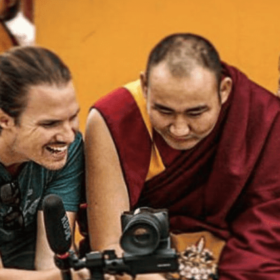 Three men, including two Buddhist monks in maroon robes and one man in a green shirt, share a joyful moment as they review their 60 seconds of production power on a camera against a bright orange background.