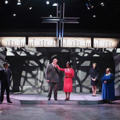 A group of actors in various costumes stand on a stage with a geometric backdrop and a large cross above, under dramatic lighting, highlighting the challenges of adaptation when translating stage plays into other forms.