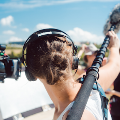 A person wearing headphones holds a boom microphone on an outdoor film set, with a camera operator and another crew member standing nearby in a sunny, rural UK landscape, capturing the action for NEEDaFIXER, a leading video production company.