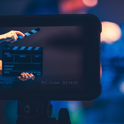 A person holds a film clapperboard in front of a camera, seen through the camera’s display screen, with a blurred blue and purple background suggesting a film production set.