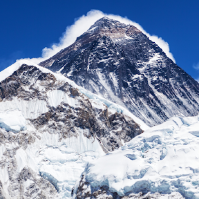Snow-covered peak of Mount Everest rises majestically against a clear blue sky, with rugged icy slopes and ridges in the foreground—a perfect subject for extreme environment photography. Wisps of clouds curl near the summit.