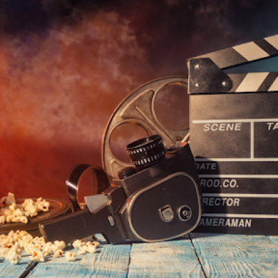A vintage movie camera, film reel, and clapperboard sit on a wooden surface with scattered popcorn and a red popcorn box—an inspiring scene for filmmakers and video production brands, all illuminated by a spotlight against a smoky, colorful backdrop.