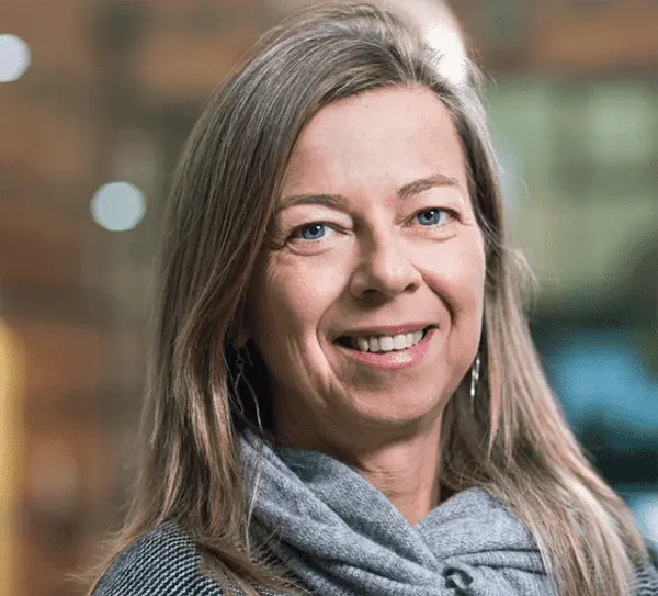 A woman with long, straight, light brown hair and blue eyes smiles at the camera. She is wearing a gray scarf and top, with a softly blurred indoor background, hinting at a Greece-based TV production setting.