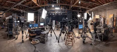 A film studio in Tirana filled with professional video cameras, lights, monitors, and equipment, set up on tripods and tables in an industrial space with exposed beams and scattered gear—perfect for filming in Tirana.