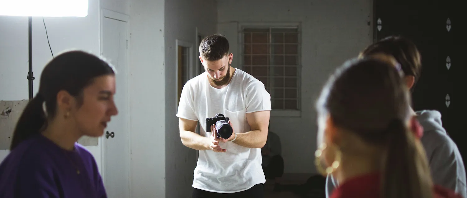 A man in a white t-shirt holds a camera and looks at the screen, while three people stand in the foreground of a dimly lit indoor film set.