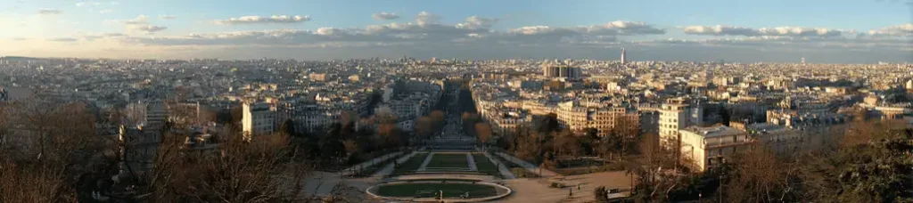 Panoramic view of Paris at sunset, showing dense city buildings, gardens, and tree-lined avenues under a partly cloudy sky—perfect for filming in Paris. The Eiffel Tower is visible in the distant right background.
