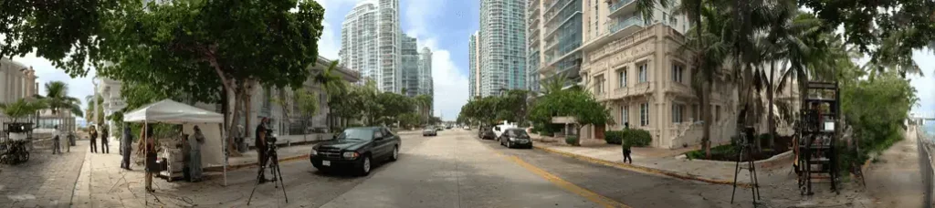 A wide city street in Miami lined with tall modern buildings and palm trees, with parked cars along the sides and some people and filming equipment visible on the sidewalks—perfect for filming in Miami.