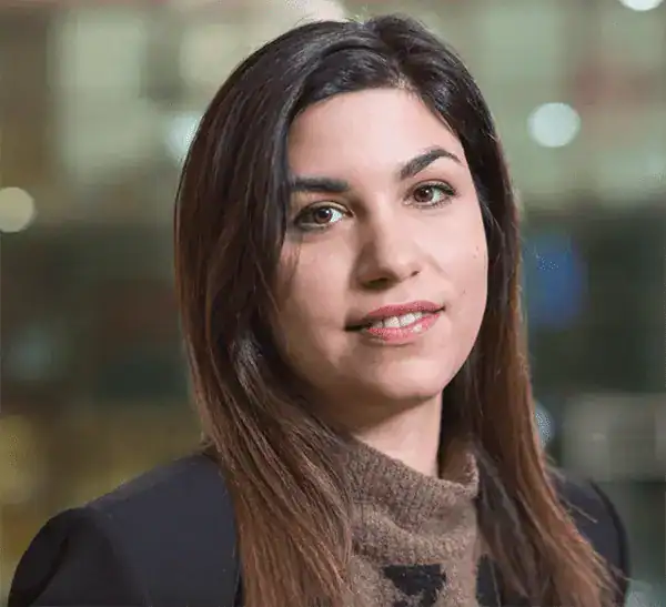 A woman with straight brown hair, dressed in a dark blazer and brown turtleneck sweater, looks at the camera and smiles softly. The blurred lights evoke a cinematic feel, reminiscent of scenes from film features set in Greece.