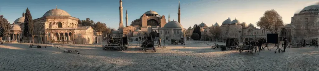 A wide panoramic view of a historic square with the Hagia Sophia in the center, surrounded by domed buildings and cobblestone pavement, captures the magic of filming in Istanbul with film equipment scattered around at sunset.