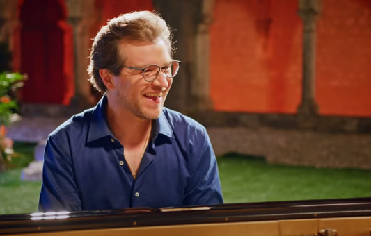 A man with glasses and a blue shirt smiles while playing piano outdoors, with stone arches and an orange wall in the background—capturing the spirit of Carnival Capriccio by Joachim Horsley.