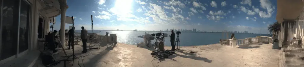 A wide shot of a film crew with cameras and equipment set up on a terrace overlooking a bay, filming in Miami, with the city skyline and blue sky with scattered clouds in the background.