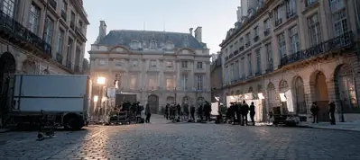 A film crew sets up equipment and lighting on a cobblestone street lined with historic buildings, filming in Paris as they prepare for a shoot in front of an ornate, classical-style building at dusk.