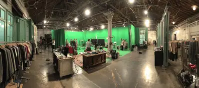 Wide view of a film studio in Paris with green screens, lighting equipment, clothing racks, costumes, and crew members preparing a set. The space features high ceilings and exposed beams, capturing the energy of filming in Paris.
