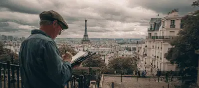 An artist wearing a cap sketches on a pad outdoors, overlooking Paris as the Eiffel Tower rises in the distance under a cloudy sky—a scene perfect for filming in Paris, surrounded by historic buildings and lush trees.