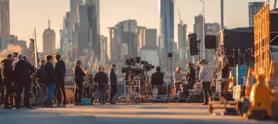 A film crew sets up equipment and cameras on an urban street with a city skyline in the background, filming in Melbourne as they work together in the early morning or late afternoon sunlight.