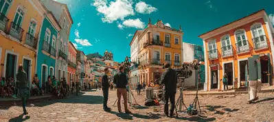 A film crew sets up cameras on a sunny cobblestone street lined with colorful colonial buildings in Rio de Janeiro, while people watch from the sidewalks as filming unfolds.