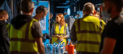 A woman in a yellow safety vest stands in the center of a busy workplace, surrounded by others in similar vests. Tables, bottles, and equipment suggest they may be filming in Barcelona.