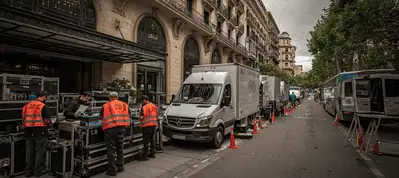 Several workers in orange vests organize equipment from trucks lined up along a city street outside a large, elegant building, suggesting preparations for filming in Barcelona.
