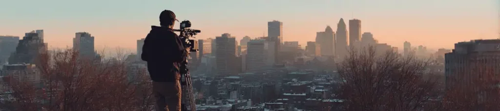 A person stands outdoors filming in Montreal with a camera on a stabilizer, overlooking the city skyline at sunset as tall buildings are silhouetted against the glowing sky.