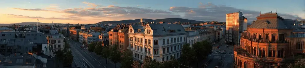 A panoramic view of Oslo at sunset, perfect for filming in Oslo, showcases historic and modern buildings, tree-lined streets, and distant hills under a sky with clouds reflecting the warm golden light.