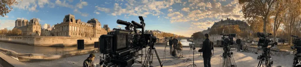A panoramic view of a riverside city scene at sunset, with several professional video cameras and crew set up in the foreground, filming in Paris amid historic buildings, a bridge, and autumn trees lining the river.